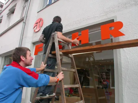 Zwei Helfer montieren auf einer Leiter die orangefarbenen Buchstaben des Schriftzugs an die Fassade