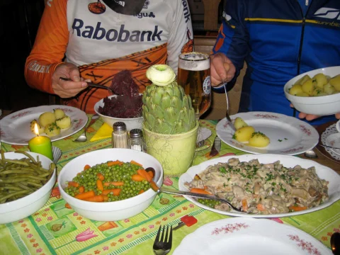 Ein gedeckter Tisch mit Kartoffeln, Gemüse und Bier, ringsum Fahrer in Trikots