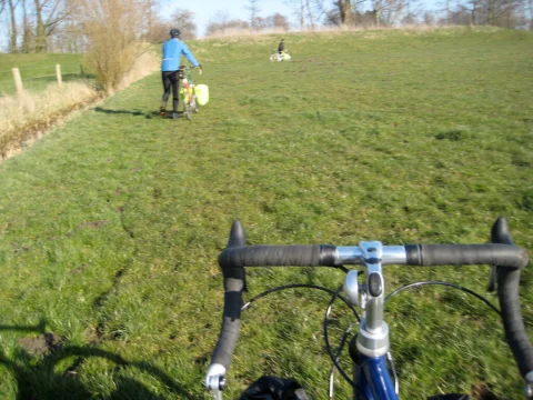 Blick über den Lenker auf einen Grasweg, in der Ferne ein Radfahrer