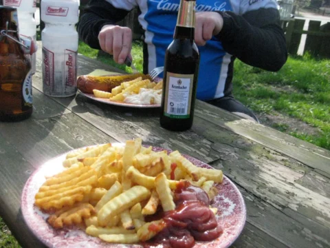 Pommes und Currywurst auf einem Pappteller auf einem Gartentisch, dahinter Radfahrende