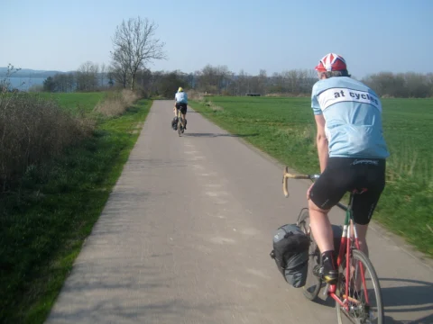 Ein Fahrer im at-Trikot auf einem Radweg zwischen Feldern, vorn weitere Fahrer