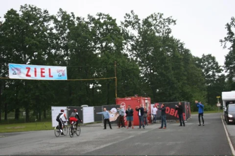 Ein Zielbanner über einer Straße, dahinter Zuschauer und ein Verpflegungsstand