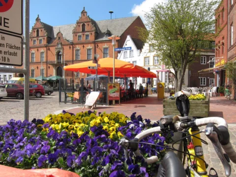 Ein Marktplatz mit Backsteinrathaus und Sonnenschirmen, vorn ein Blumenbeet und ein Lenker