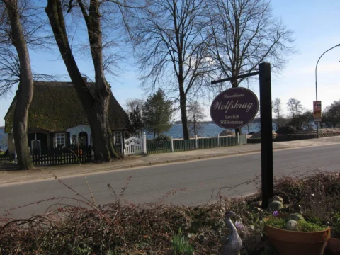 Eine Landstraße vor einem reetgedeckten Gasthaus, dahinter das Wasser
