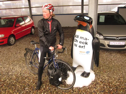 Ein Fahrer neben einer Pinguinfigur mit der Aufschrift „Klima-Check hier!“