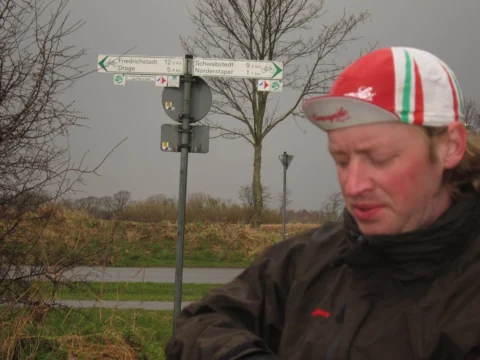 Ein Fahrer in Regenjacke an einer Kreuzung, dahinter ein Wegweiser