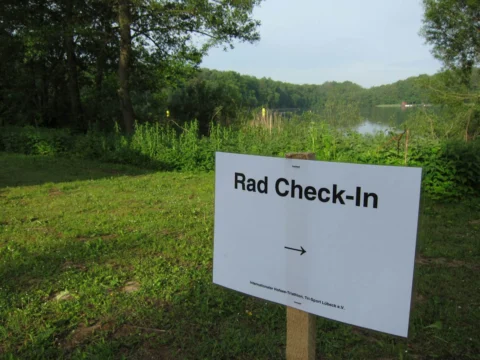 Ein Schild „Rad Check-In“ mit Pfeil auf einer Wiese am See