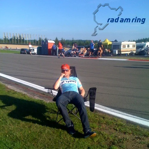 Ein Fahrer im at-Trikot sitzt im Campingstuhl am Rand der Rennstrecke, dahinter das Nürburgring-Logo