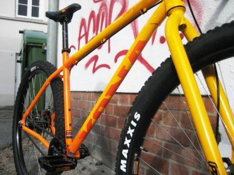 Ein orange-gelbes Kona Unit lehnt an einer Backsteinwand mit Graffiti