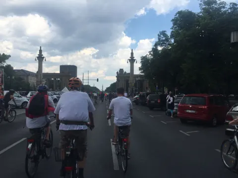 Radfahrende auf einer Allee zwischen Säulen, rechts stehende Autos
