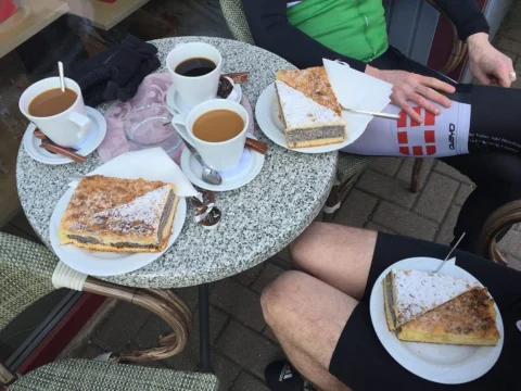 Kaffee und Kuchenstücke auf einem Bistrotisch, ringsum Radfahrende in Trikots
