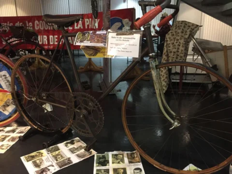 Ein historisches Fahrrad in einer Ausstellung, dahinter alte Rennplakate