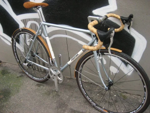 Ein silbernes Rennrad mit braunem Lenkerband und Holzschutzblechen an einer Graffitiwand