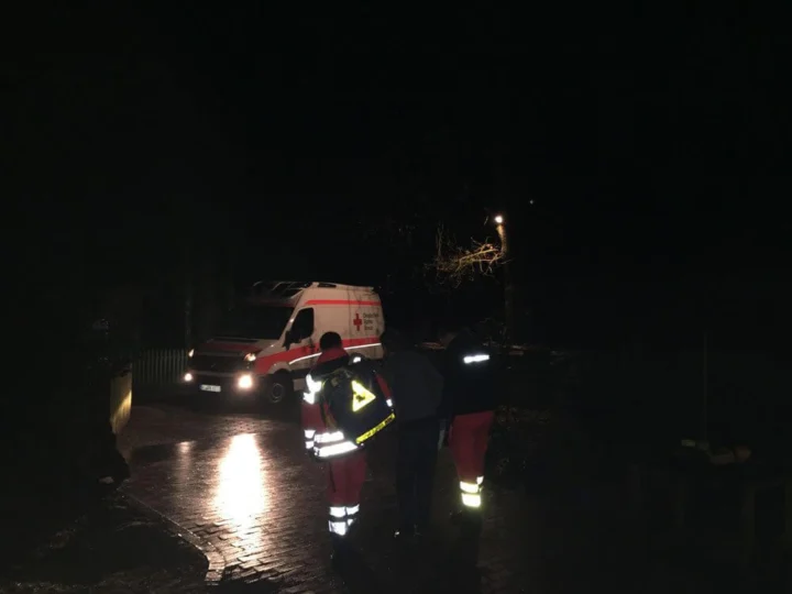 Ein Einsatzfahrzeug bei Nacht auf einer nassen Straße, im Licht der Scheinwerfer eine Person in Warnkleidung