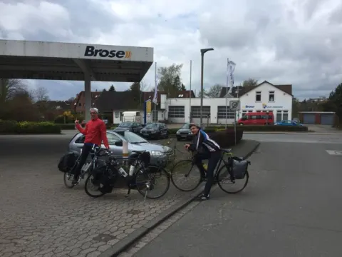 Zwei Radfahrer mit bepackten Rädern auf einem Tankstellenvorplatz