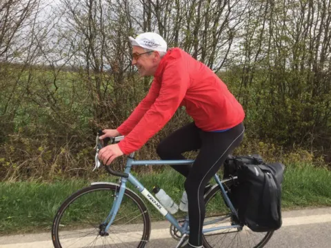Ein Fahrer in roter Jacke auf einem Reiserad mit Packtasche, dahinter eine Hecke