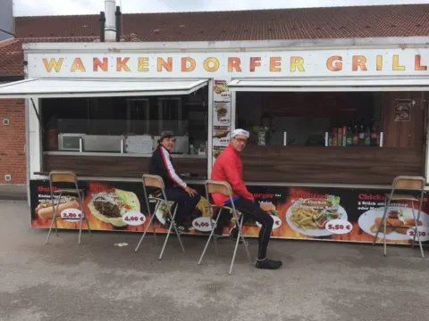 Zwei Radfahrer sitzen vor einem Imbisswagen mit der Aufschrift „Wankendorfer Grill“