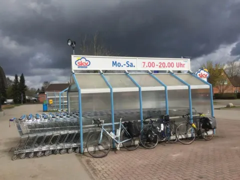 Zwei bepackte Räder stehen vor Einkaufswagen an einem Supermarkt unter dunklen Wolken