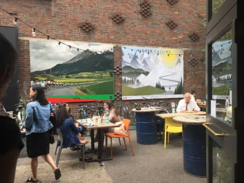 Gäste an Tischen in einem Café, an der Backsteinwand hängen große Fotoplakate