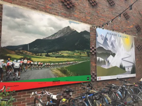 Zwei große Plakate an einer Backsteinwand: ein Feld auf einer Passstraße und eine Berggrafik