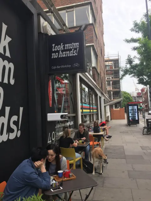 Die Fassade eines Cafés mit großem Schriftzug, davor sitzen Gäste an Tischen