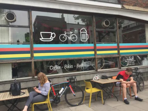 Eine Fensterfront mit den Symbolen Kaffee, Fahrrad und Bier, davor Gäste und abgestellte Räder