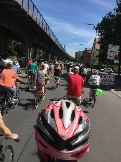 Ein Pulk Radfahrender unter einer Hochbahnbrücke, im Vordergrund ein rosa Helm