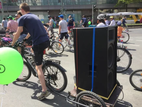Ein Radfahrer transportiert einen großen Lautsprecher auf dem Gepäckträger, daneben ein grüner Luftballon