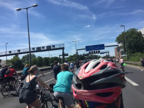 Radfahrende auf der Autobahn unter einer Schilderbrücke, im Vordergrund ein roter Helm