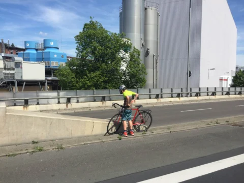 Ein einzelner Fahrer im gelben Trikot auf der leeren Fahrbahn, dahinter Industriebauten