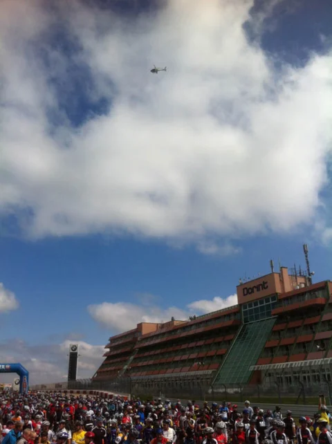 Blick über das wartende Starterfeld zur Tribüne, darüber ein Hubschrauber am Wolkenhimmel