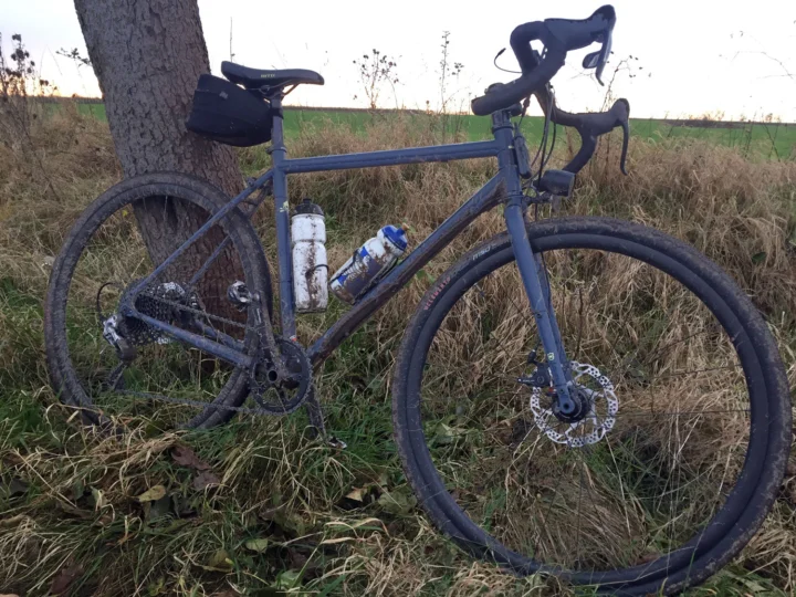Ein graues Kona Rove ST mit Satteltasche lehnt an einem Baum im Feld