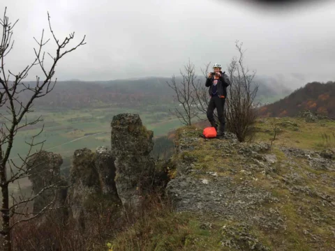 Ein Radfahrer steht auf einer Felskuppe über einem nebligen Tal, daneben liegt sein Rad