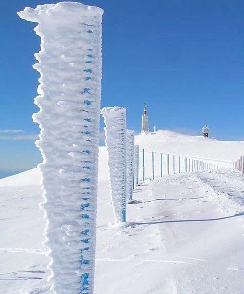 Vom Eis überzogene Zaunpfähle auf einem verschneiten Gipfel, im Hintergrund der Wetterturm