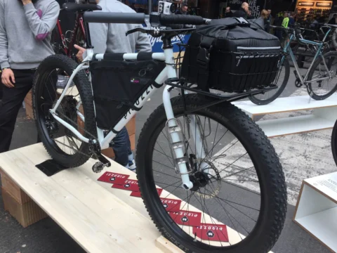 Ein weißes Rad mit stolliger Bereifung, Frontgepäckträger und Tasche auf einem Podest