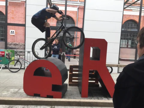 Ein Fahrer springt mit dem Rad über eine rote Buchstabenskulptur vor einer Backsteinhalle