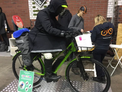 Ein Besucher in dicker Winterjacke sitzt auf einem grünen Rad, daneben Standpersonal