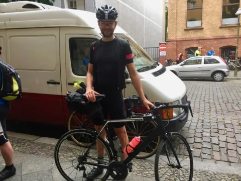 Ein Fahrer steht mit bepacktem Rennrad vor einem alten Transporter auf Kopfsteinpflaster
