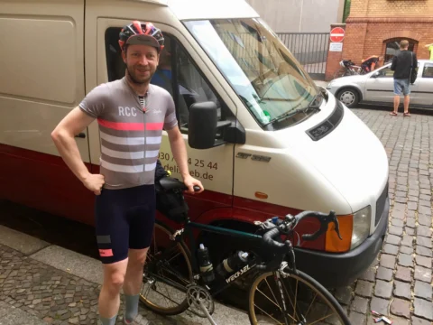 Ein Fahrer im gestreiften Trikot steht neben seinem Rad vor demselben Transporter