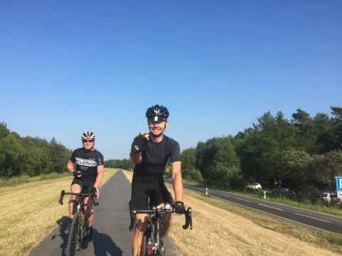 Zwei Fahrer nebeneinander auf einem Radweg unter blauem Himmel, einer winkt in die Kamera