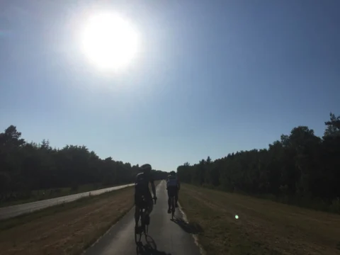 Zwei Fahrer als Silhouetten auf einem geraden Weg, darüber die tief stehende Sonne im Dunst