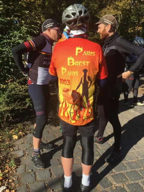 Drei Teilnehmende im Gespräch, einer trägt ein oranges Paris-Brest-Paris-Trikot von 2003