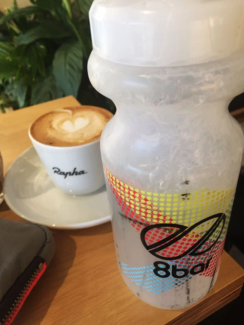 Eine Trinkflasche voller Eis neben einem Cappuccino auf einem Cafétisch