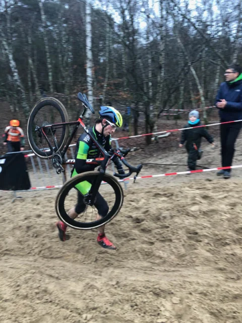 Ein Fahrer trägt sein Rad durch die Sandpassage, dahinter Absperrband und Publikum