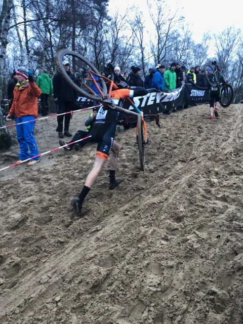 Ein Fahrer läuft mit geschultertem Rad die Sandwand hinauf, oben stehen Zuschauer