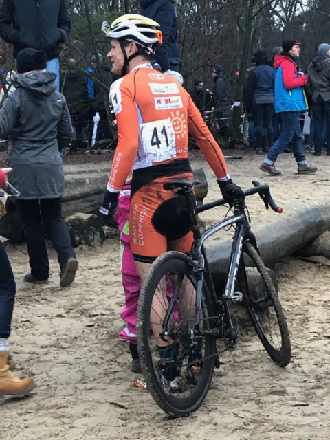 Eine Fahrerin im orangefarbenen Trikot mit Startnummer 41 schultert ihr Rad neben Zuschauern