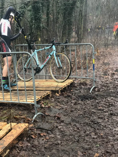 Ein Fahrer schultert sein Rad über eine Holzrampe im Matsch