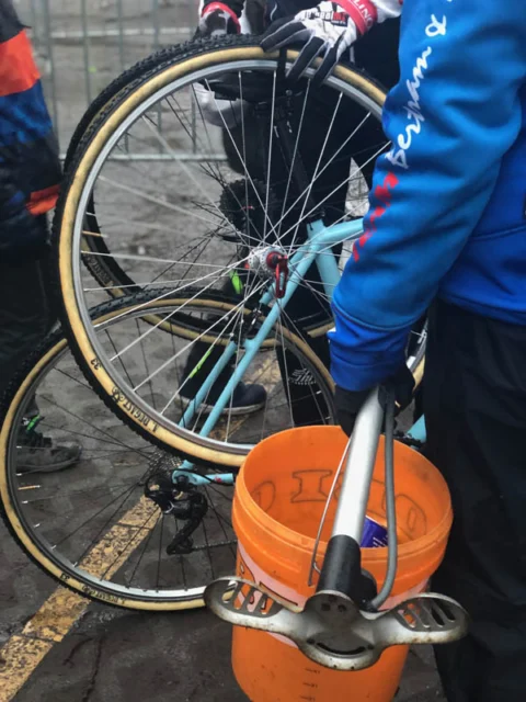 Ein Helfer wäscht ein Rad mit dem Schlauch über einem orangefarbenen Eimer