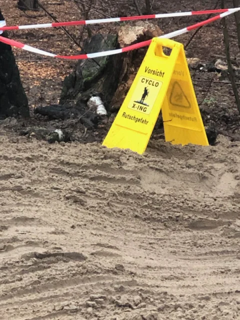 Ein gelbes Warnschild „Vorsicht Cyclo X-ing, Rutschgefahr“ steht in einer aufgewühlten Sandpassage