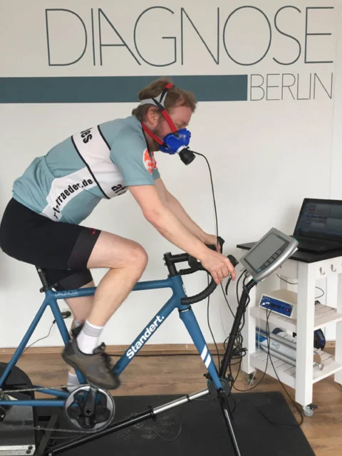 Ein Fahrer auf dem Ergometer mit Atemmaske bei der Leistungsdiagnostik, daneben ein Messgerät